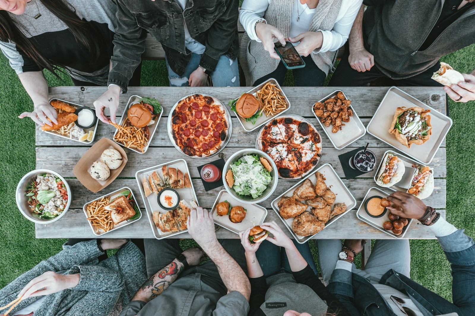 Een lange houten tafel van bovenaf vol gerechten, met handen die van alle kanten iets pakken