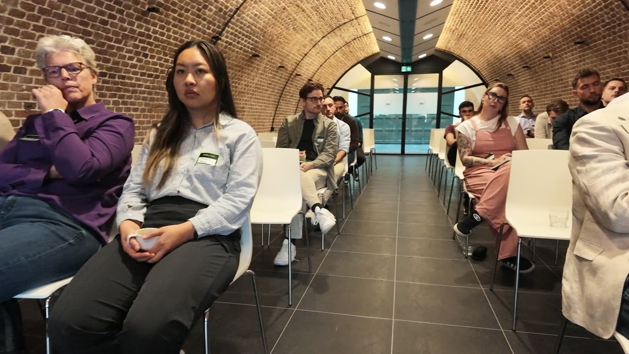 Gasten in een kleine zaal bij Finance Unlocked, met een camera die meekijkt